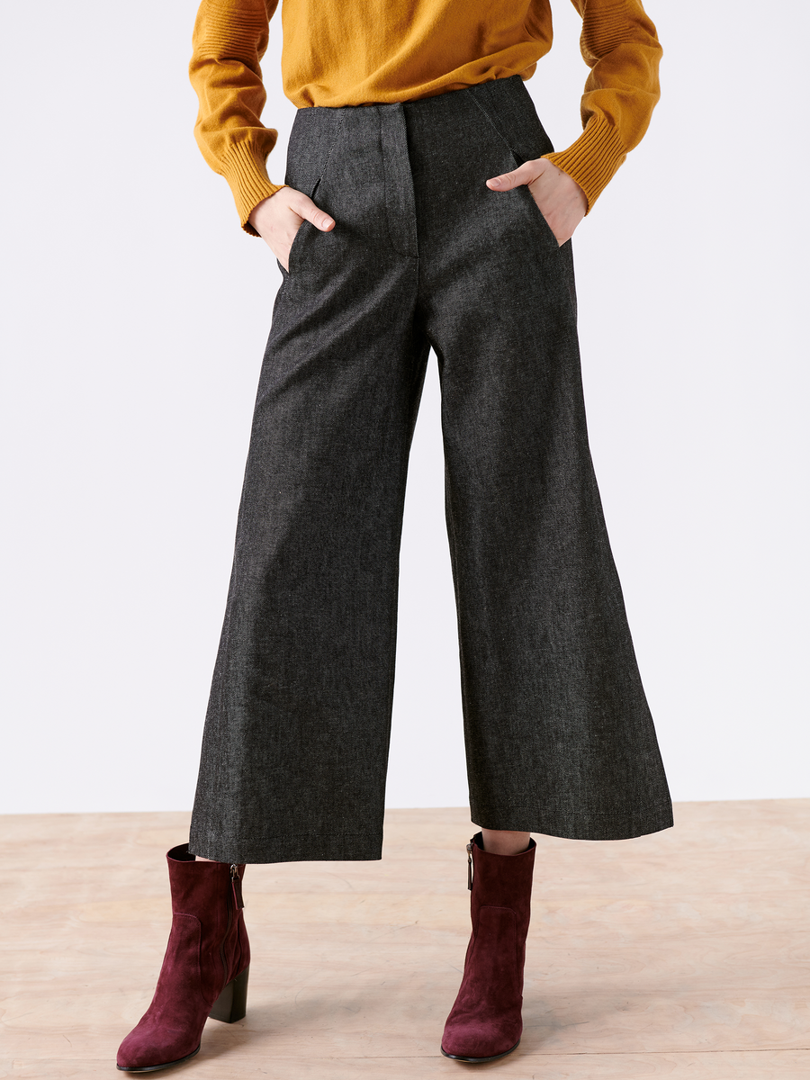 From the shoulders down, a person is seen in a mustard sweater with hands in the pockets of Zero + Maria Cornejo's Cropped Fin Pant made from recycled cotton, paired with burgundy ankle boots with heels, standing on a light wooden floor.