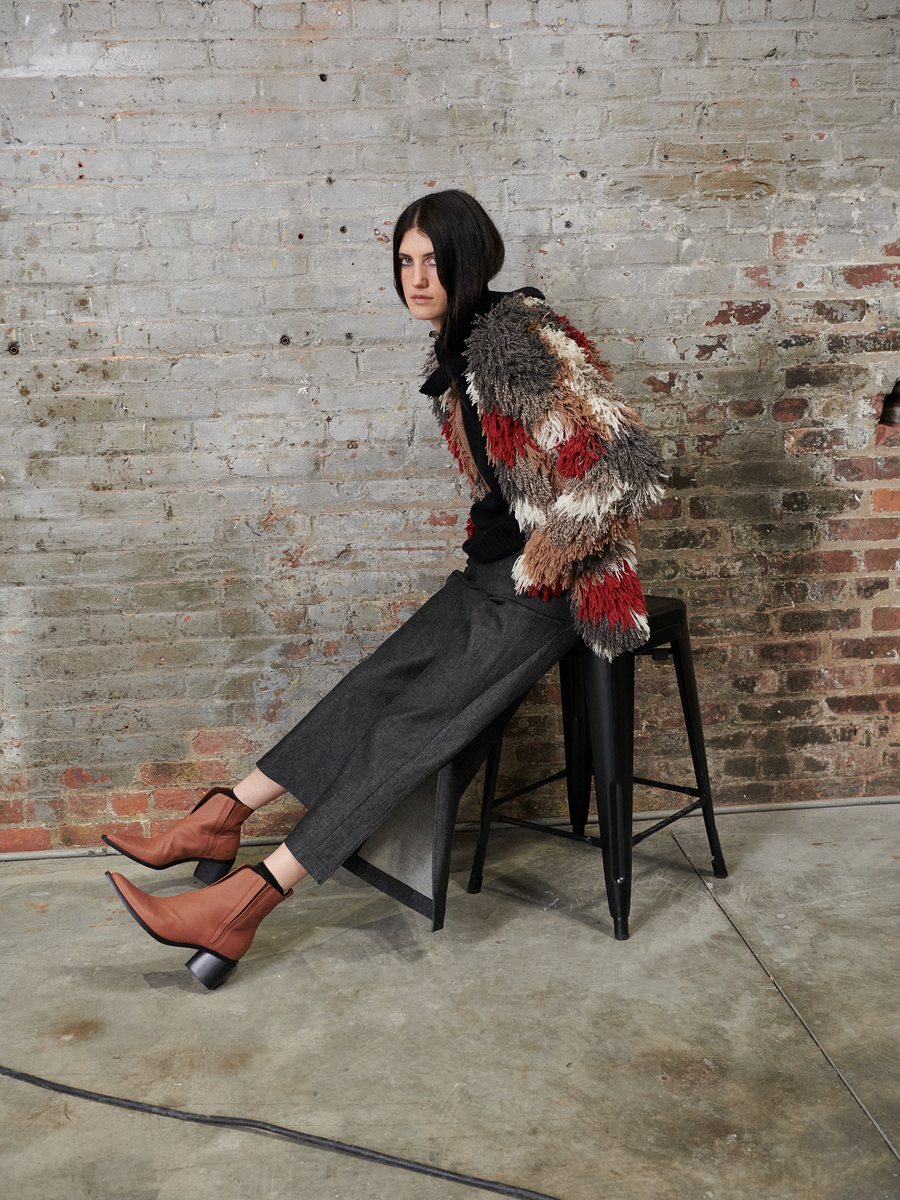 A person with long dark hair leans against a brick wall on a black stool, wearing a colorful textured jacket and brown ankle boots. They sport the Cropped Fin Pant by Zero + Maria Cornejo, made from recycled cotton with EarthColors technology, adding to the rustic, industrial vibe.