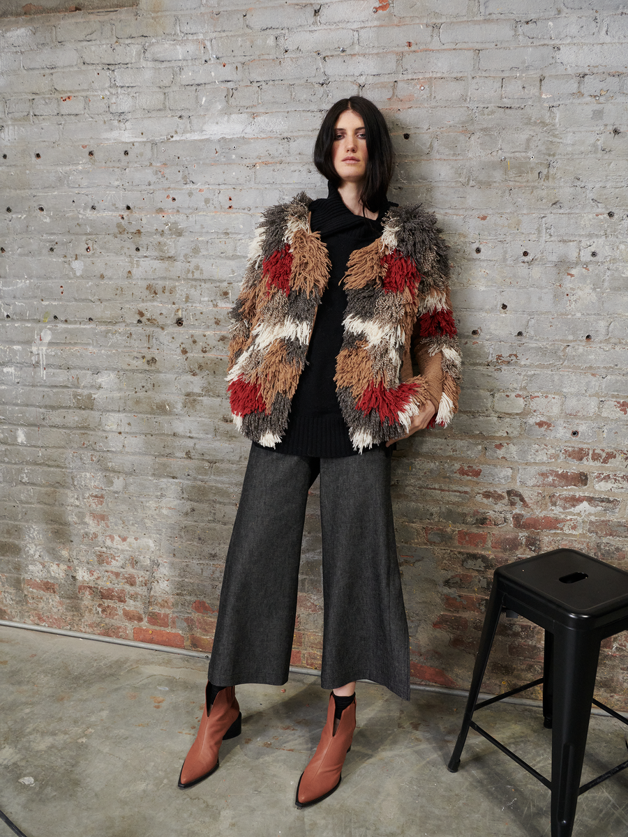 A person leans on a black stool, wearing a multicolored textured jacket, a black top, and Zero + Maria Cornejo's Cropped Fin Pant in wide-legged gray denim with EarthColors technology. Brown ankle boots enhance the look. With short dark hair and hands in their pockets, they stand against a brick wall.