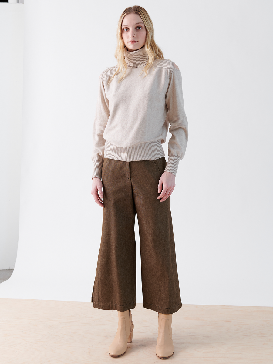 A person with long blonde hair stands in a white studio wearing a beige turtleneck, Zero + Maria Cornejo's Cropped Fin Pant made from recycled cotton, and beige ankle boots while facing the camera neutrally.