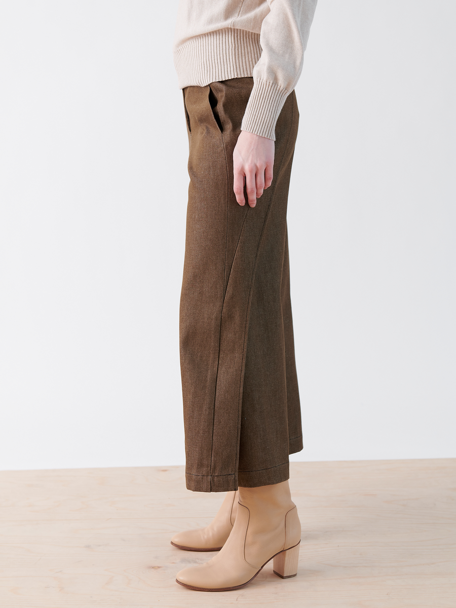 A person is wearing Zero + Maria Cornejo's Cropped Fin Pant in brown, made with Archroma’s EarthColors® technology, paired with a light beige sweater and tan ankle boots. They are standing on a light wood floor in front of a white wall, captured from the side highlighting their lower half.