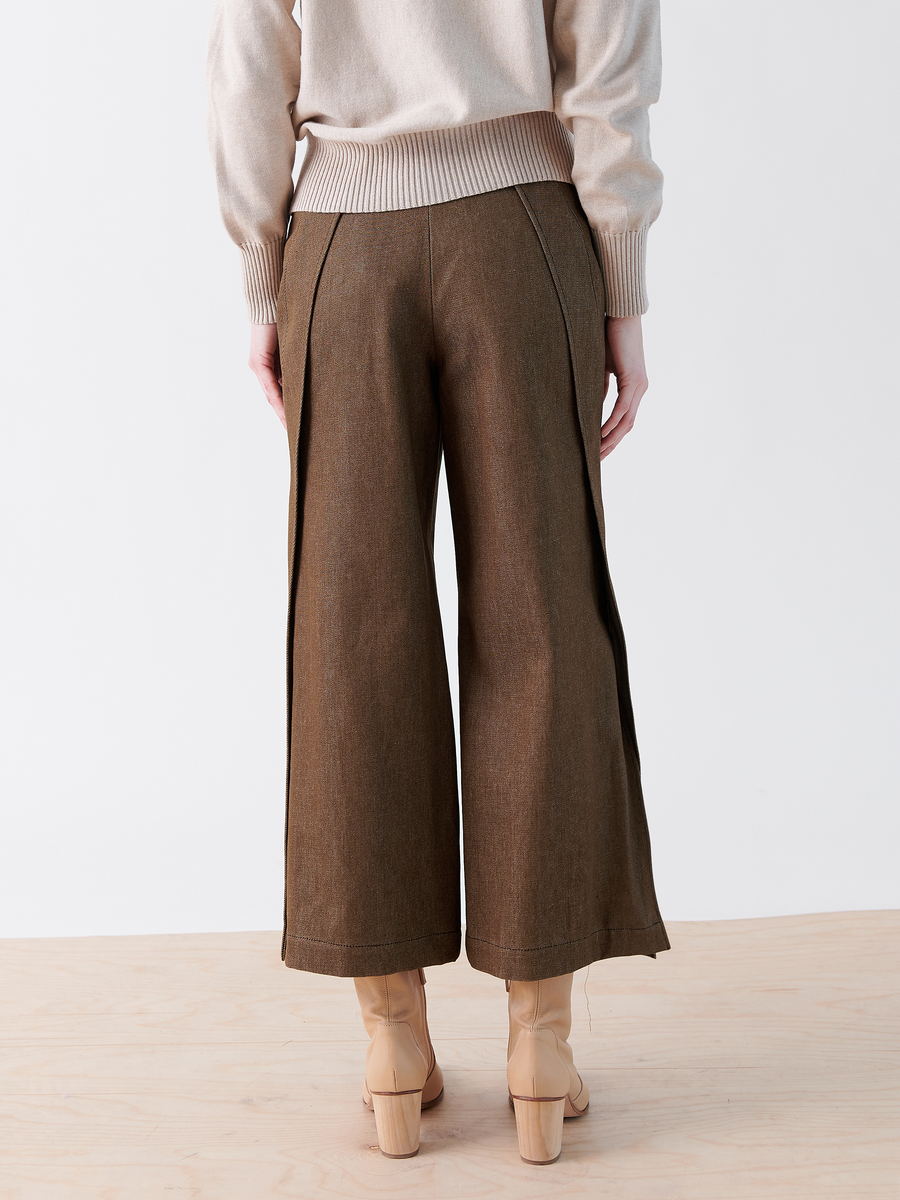 A person is seen from the back wearing Zero + Maria Cornejo's Cropped Fin Pant, featuring wide-leg design and modern side panels like traceable denim, paired with a light beige sweater and beige heeled boots. The floor is wooden and the background is plain white.