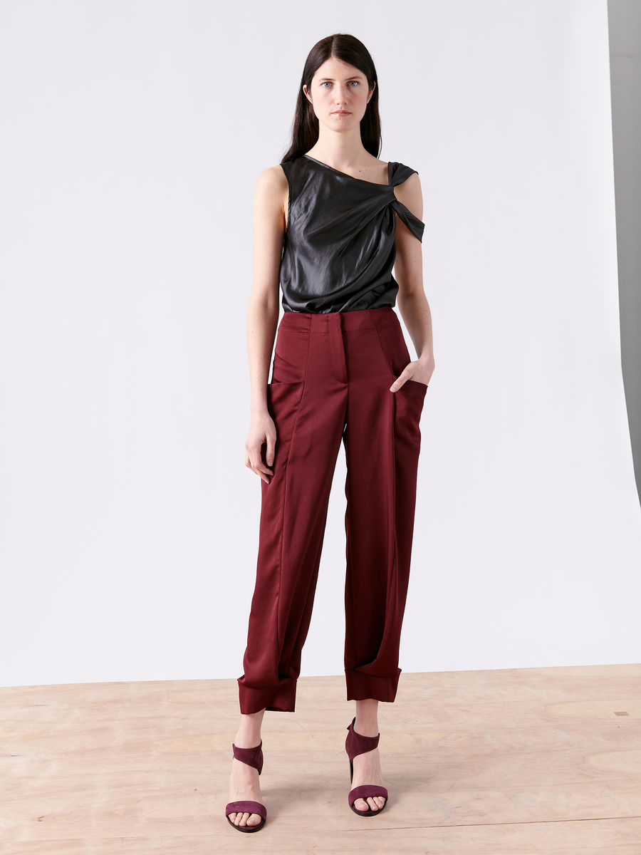 A woman stands on a wooden floor against a plain white background wearing the asymmetrical black leather top and Akeo Jodhpur high-waisted burgundy drape pants by Zero + Maria Cornejo, crafted in New York from 100% recycled fabric. Her left hand rests in her pocket, paired with matching burgundy heels.