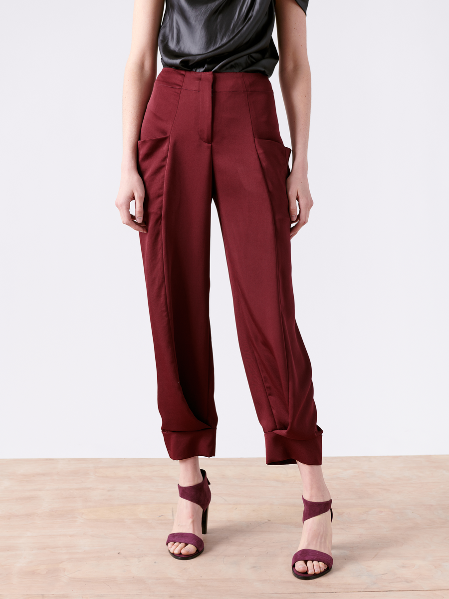 A person poses on a wooden floor wearing Zero + Maria Cornejo's Akeo Jodhpur trousers, crafted from 100% recycled fabric in burgundy, paired with matching heels and a dark shirt. The image highlights this sustainable fashion ensemble made in New York.