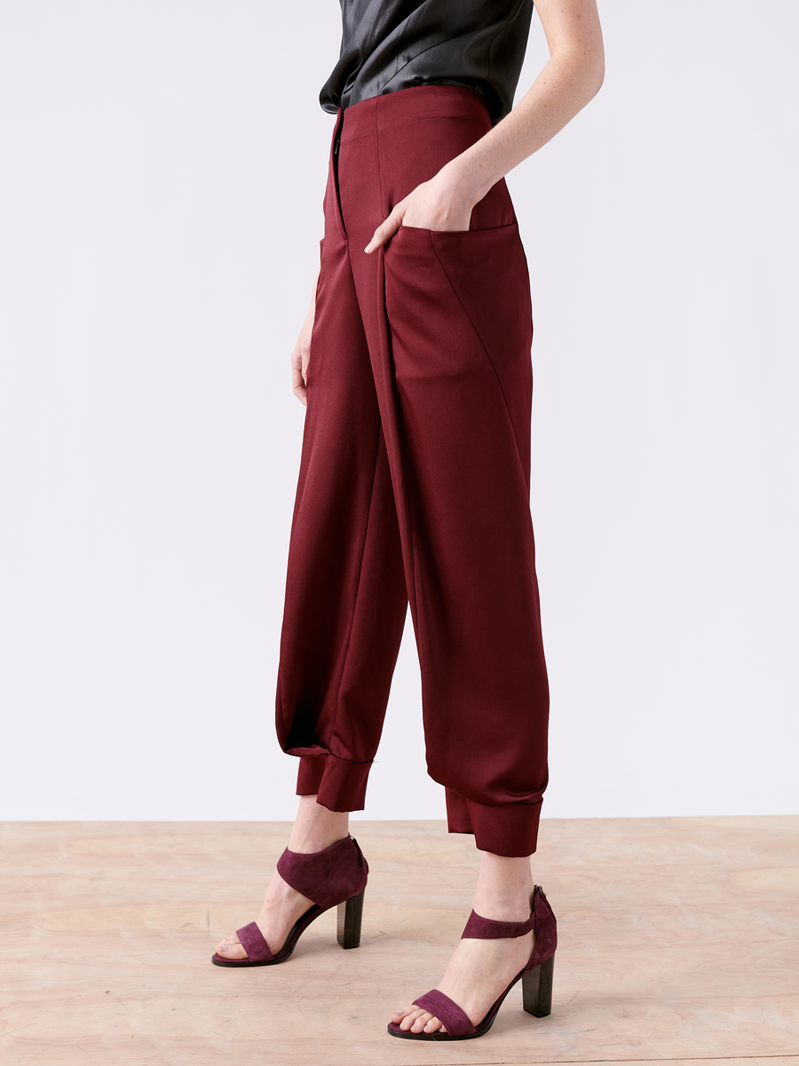 Against a plain white background, someone stands on a wooden floor wearing Zero + Maria Cornejo's Akeo Jodhpur in maroon, made from 100% recycled fabric with large pockets. They pair these harem pants with matching high-heeled sandals and a dark top for a minimalist, sustainable look.