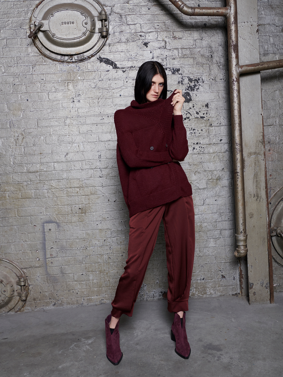 A person with short dark hair stands against an industrial-style brick wall, wearing a cozy maroon turtleneck sweater and recycled drape pants made from 100% recycled fabric. The Akeo Jodhpur ankle boots by Zero + Maria Cornejo complete their sustainable look, with metal pipes enhancing the backdrop.