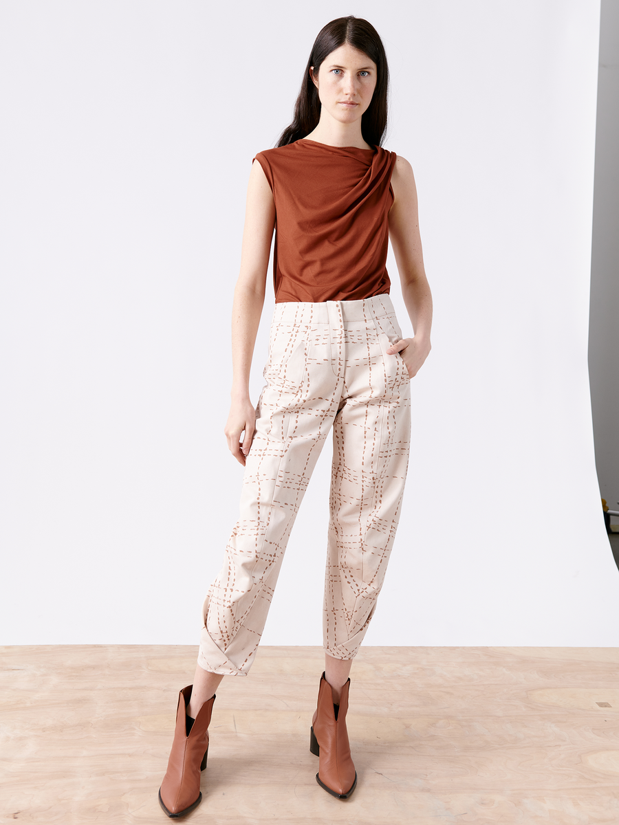 A person stands confidently on a wooden floor, wearing the rust-colored sleeveless top with Takeo Pant by Zero + Maria Cornejo, featuring cream organic cotton and a geometric pattern. Brown ankle boots complete the look against a plain white background.