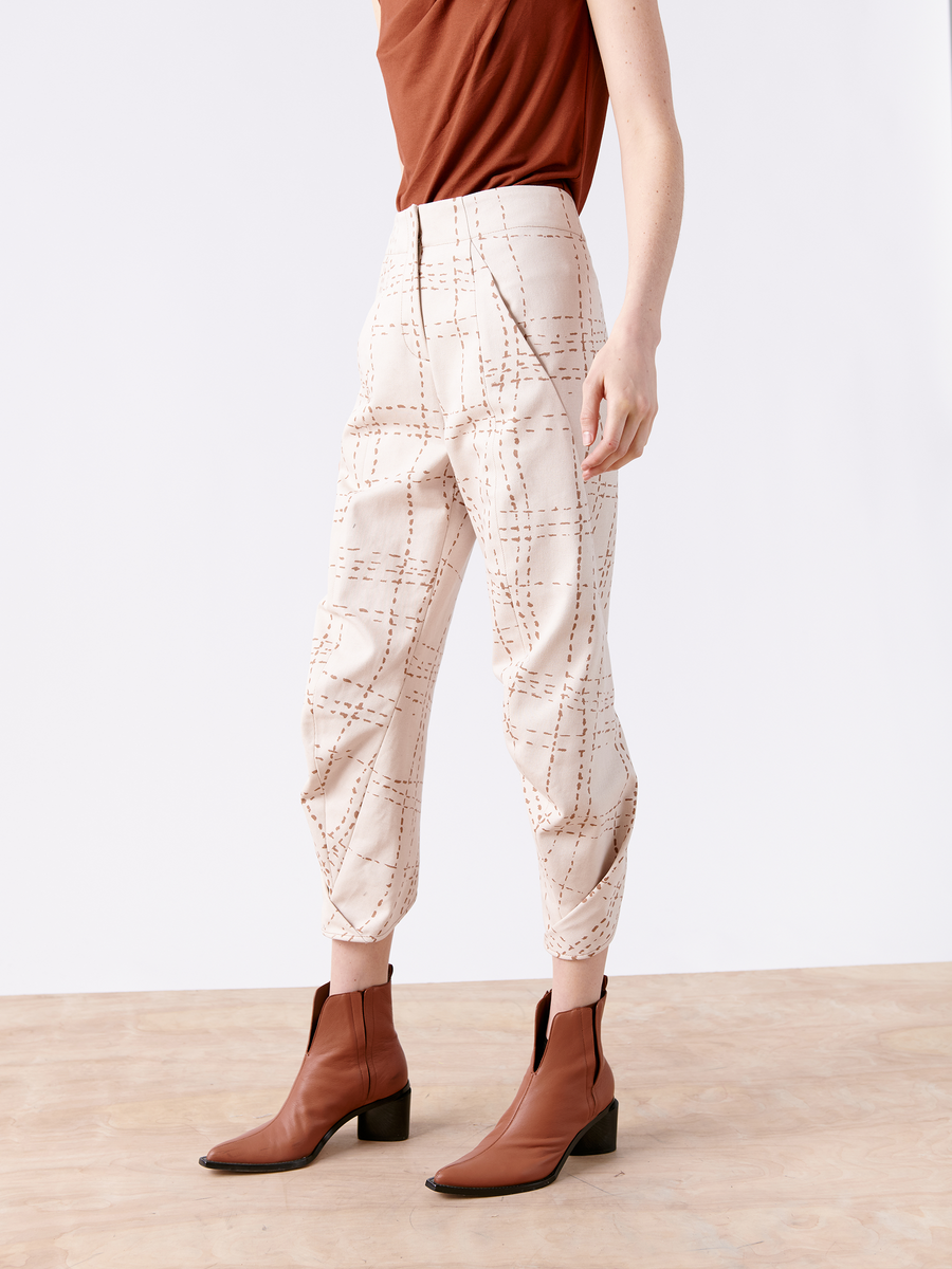 A person is standing on a wooden floor wearing a sleeveless brown top, the Takeo Pant from Zero + Maria Cornejo's Final Sale collection, and brown ankle boots. The plain white background highlights their elegant draped silhouette.