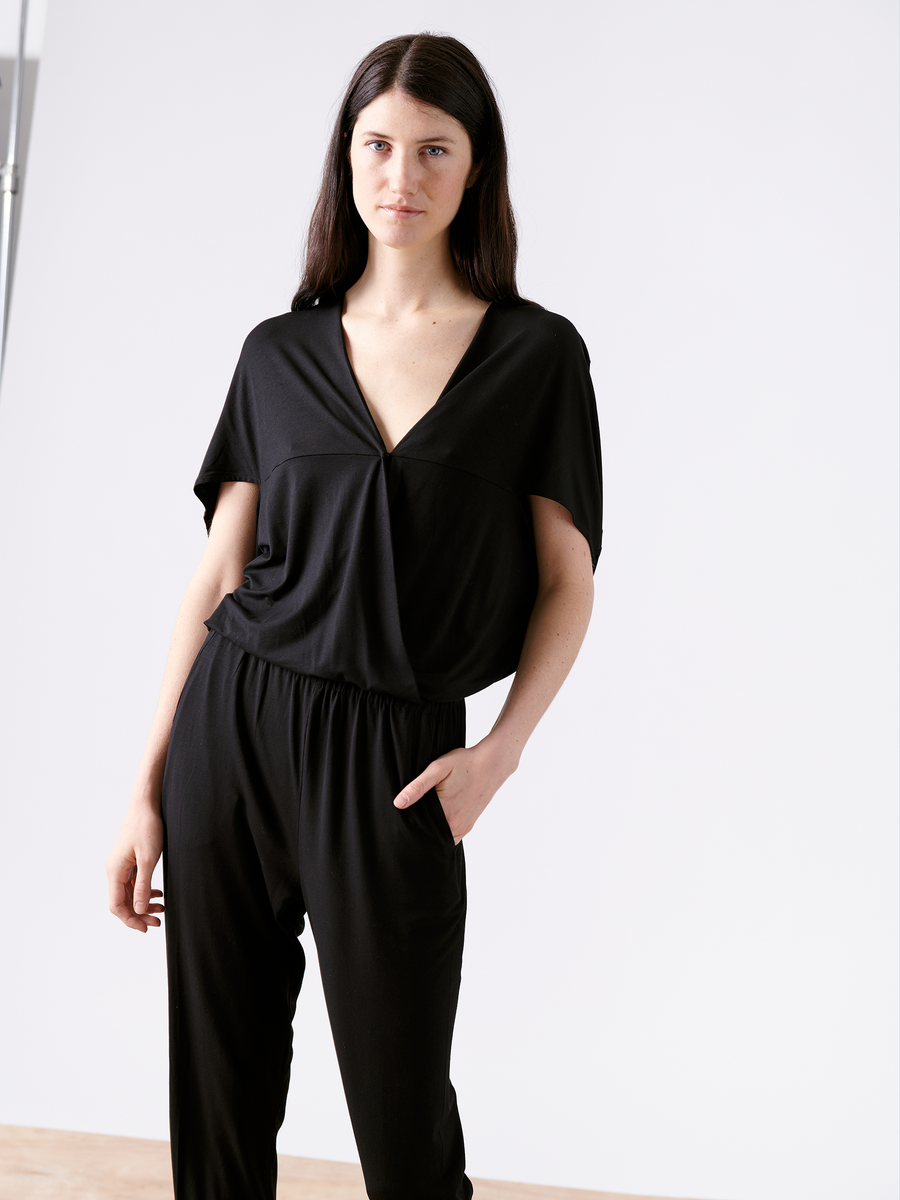 A person with long dark hair stands against a plain white background, wearing the Hex Jumpsuit by Zero + Maria Cornejo. This sleek black jumpsuit showcases an elastic waist, flattering V-neck, hands-in-pockets style with a loose-fitting top and tapered pants.