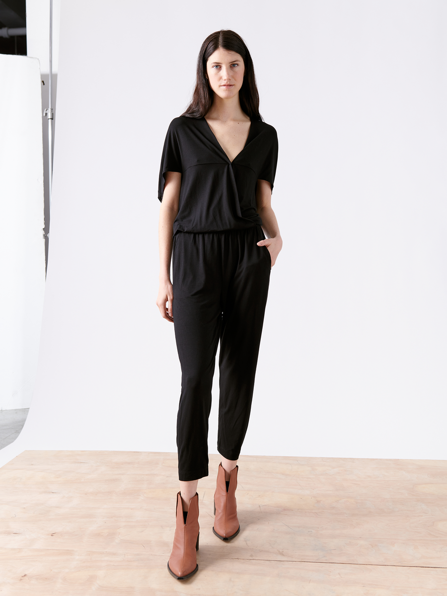 A person with long dark hair stands on a wooden floor wearing a Hex Jumpsuit by Zero + Maria Cornejo, featuring an elastic waist. They pair it with tan ankle boots and have their right hand in their pocket against a plain white and gray backdrop.