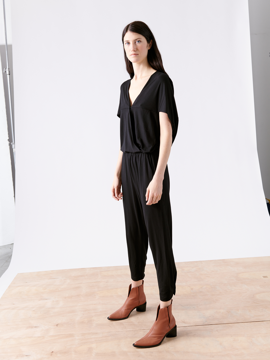 A person with long dark hair dons the Zero + Maria Cornejo Hex Jumpsuit, a black V-neck with an elastic waist, paired with brown ankle boots, standing on a wooden platform against a white backdrop.