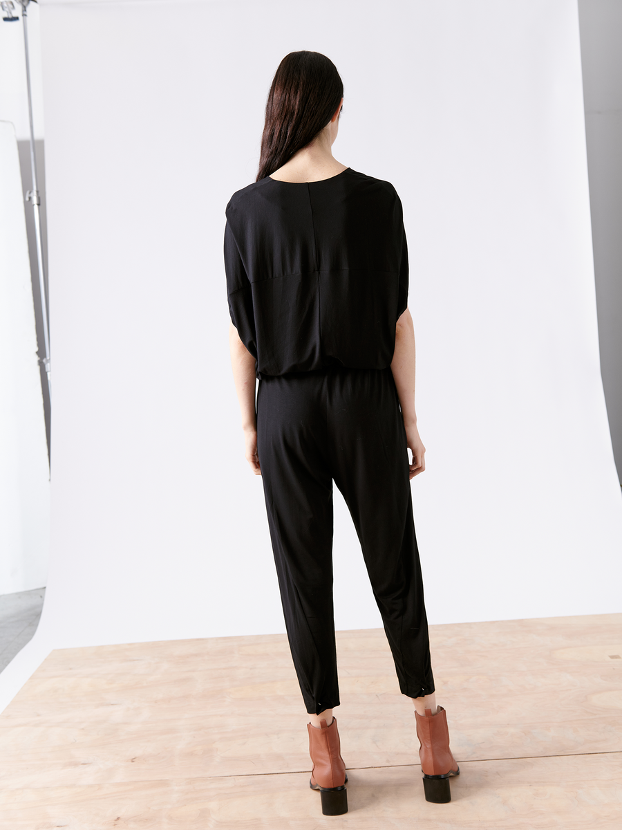 A person with long dark hair stands on a wooden floor, facing away from the camera. They're wearing the Zero + Maria Cornejo Hex Jumpsuit - Final Sale, featuring short sleeves and an elastic waist, paired with brown ankle boots. The background is a light-colored studio backdrop.