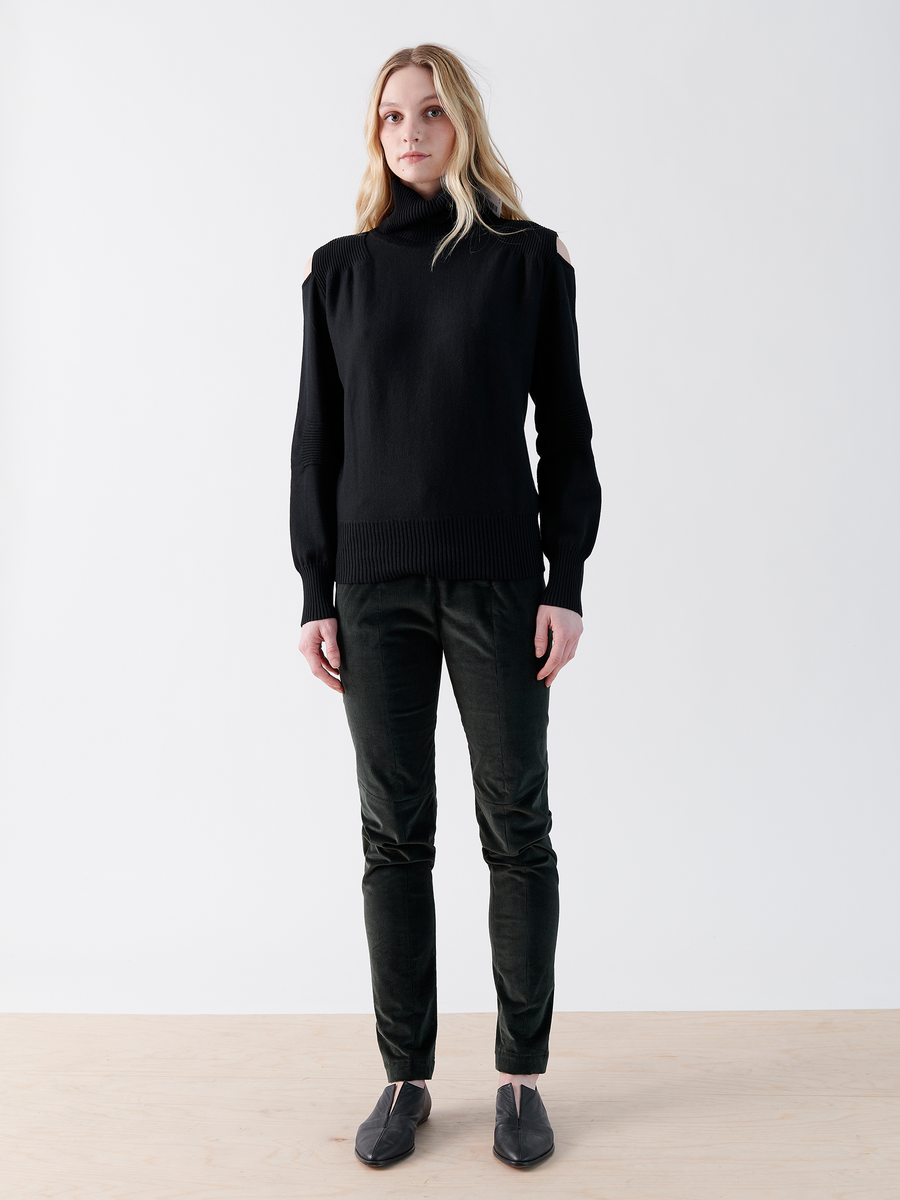 A person with long blonde hair faces forward, wearing a black turtleneck sweater with shoulder cutouts, Zero + Maria Cornejo's Slim Tala Trouser - Final Sale featuring an elastic waist, and gray flat shoes. They stand on a light wood floor against a plain white background.