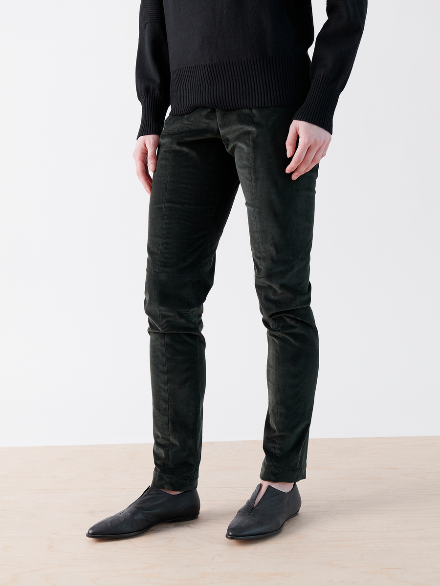 A person is wearing a black sweater, Slim Tala Trousers in dark green velvet by Zero + Maria Cornejo, and black slip-on shoes, standing against a white background.