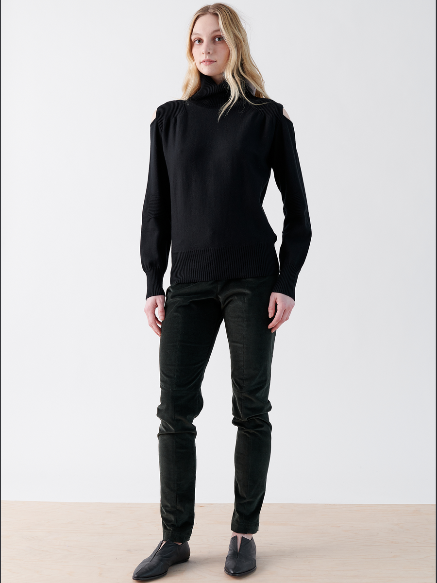 A woman with long blonde hair stands against a plain white background, wearing a black turtleneck sweater and Zero + Maria Cornejo's Slim Tala Trouser - Final Sale that complements her dark shoes. Her face holds a neutral expression.