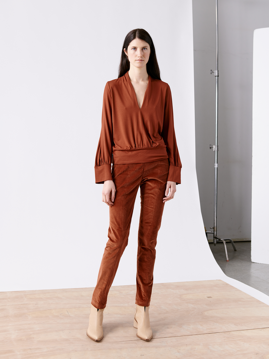 A person with long dark hair stands in a studio wearing a rust-colored wrap-top and the Slim Tala Trouser by Zero + Maria Cornejo, paired with beige ankle boots. The plain background shows some studio equipment.