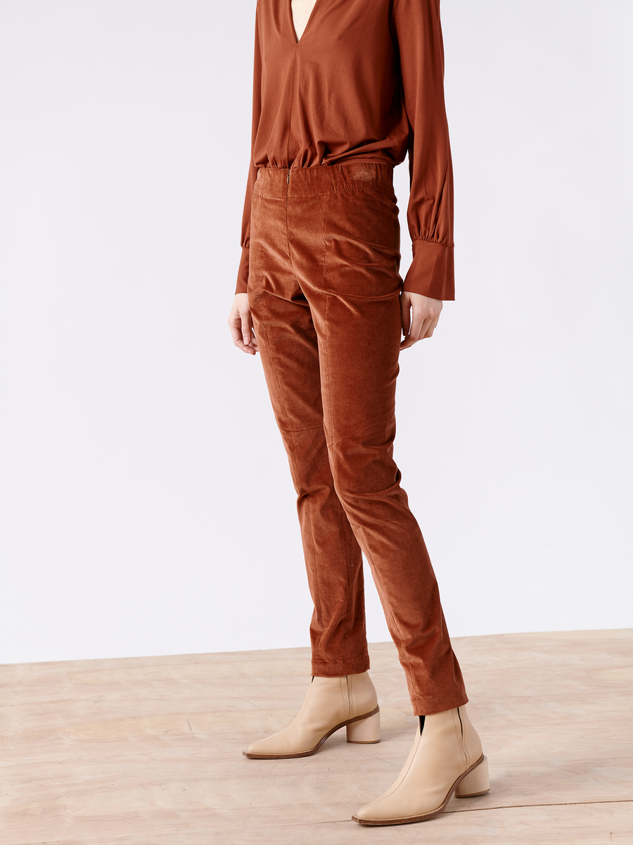 A person wearing Zero + Maria Cornejo's Slim Tala Trouser in rust, featuring an elastic waist and paired with beige ankle boots, stands on a wooden floor. Their face is unseen.