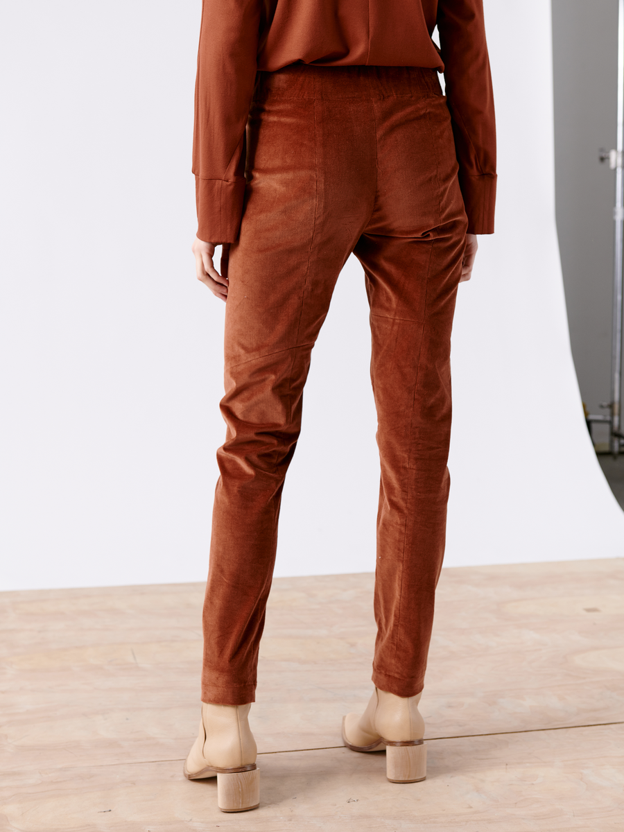 A person wears the Slim Tala Trouser by Zero + Maria Cornejo in rust-colored cotton corduroy, featuring an elastic waist, paired with a matching long-sleeve shirt. They stand on a wooden floor against a white background, sporting beige block-heeled shoes.