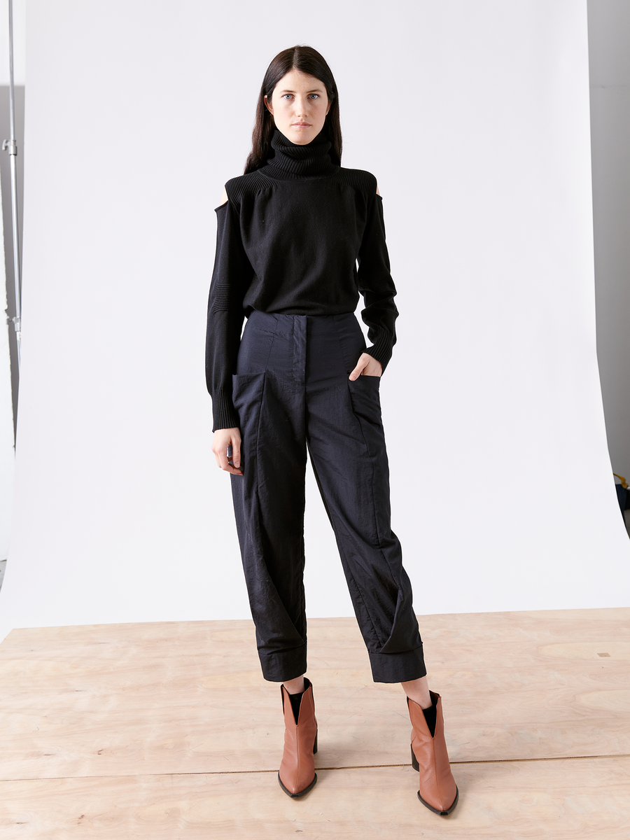 A person stands on a wooden platform against a white backdrop, wearing a black turtleneck and tech taffeta pants with pockets. Their Akeo Jodhpur boots by Zero + Maria Cornejo complement the sleek ensemble as their right hand rests in a pocket, while long dark hair cascades down.