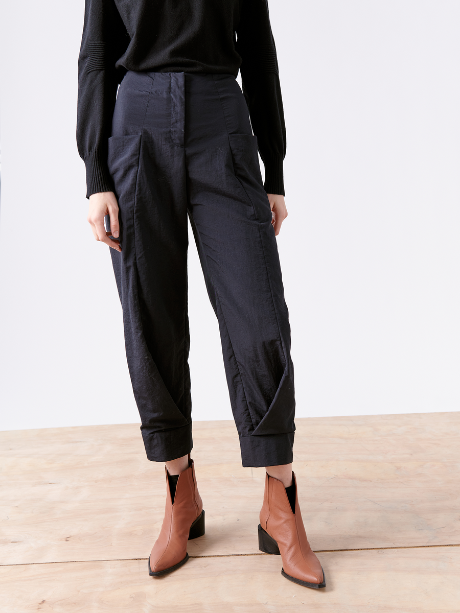 A person is wearing cuffed, loose-fitting black tech taffeta pants and a black sweater with the Akeo Jodhpur by Zero + Maria Cornejo, showcasing brown ankle boots with geometric cutout details against a plain background, emphasizing the sleek silhouette formed by the lightweight fabric.