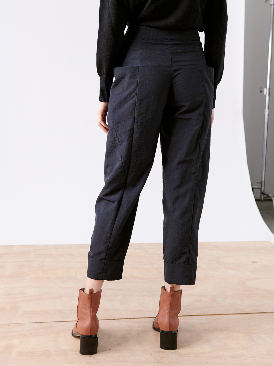 Seen from the rear, a person wears Zero + Maria Cornejo's Akeo Jodhpur, black high-waisted cropped pants made of lightweight fabric, paired with brown ankle boots. They stand on a wooden floor against a white background.