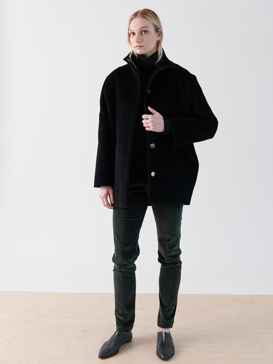 In a minimalist space, a person wears Zero + Maria Cornejo's Long-Sleeved Bea Parka in black, matched with dark pants and gray shoes. The setting features a plain white wall and light wood floor.