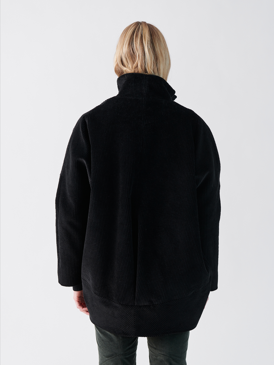 A person with blonde hair wears a black Long-Sleeved Bea Parka from Zero + Maria Cornejo and dark green, wide-wale cotton corduroy pants against a light grey background.