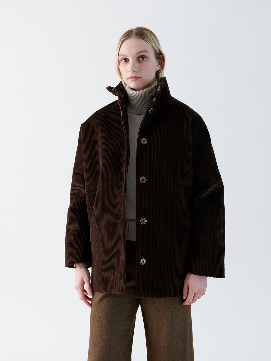 Against a white background, a person wears the Long-Sleeved Bea Parka by Zero + Maria Cornejo, with visible buttons and high collar in dark brown corduroy contrasted by light brown trousers. Shoulder-length blonde hair frames their face above a beige turtleneck sweater.