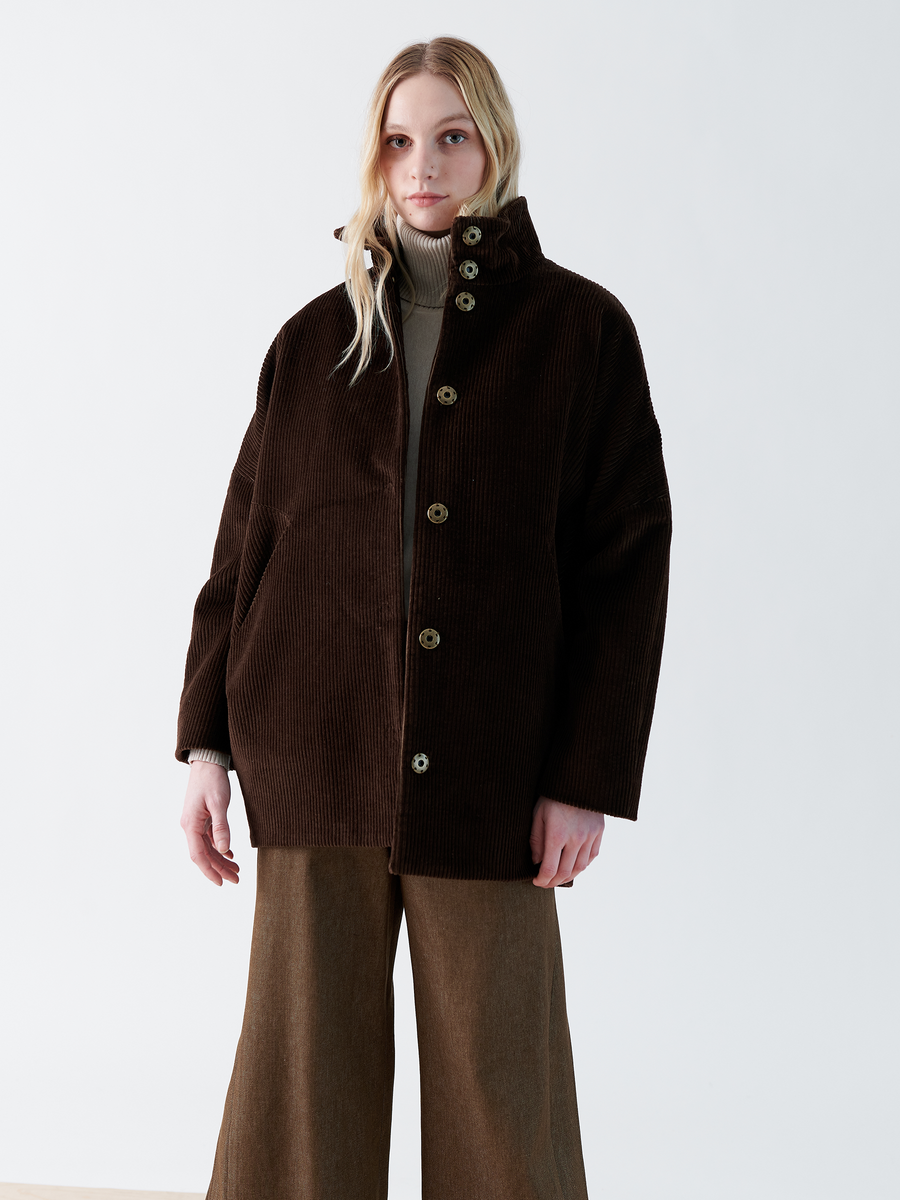 A person with long, light hair wears the Zero + Maria Cornejo Long-Sleeved Bea Parka in dark brown corduroy and brown wide-leg pants. They pair it with a light neutral top against a plain white background. The parka features large buttons and a high funnel collar.