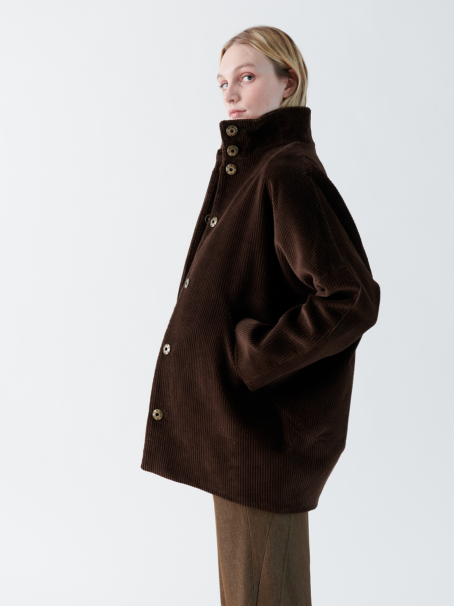 A person in a dark brown Long-Sleeved Bea Parka by Zero + Maria Cornejo, featuring wide-wale corduroy with a high funnel collar and brown buttons, gazes to the side against a simple, light background.