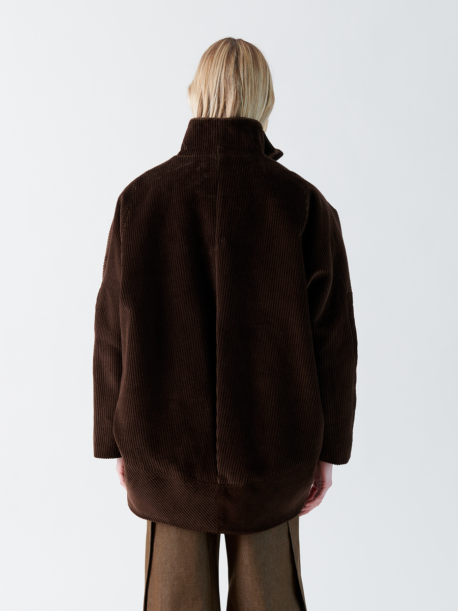 A person with shoulder-length blonde hair wears the Zero + Maria Cornejo Long-Sleeved Bea Parka in dark brown wide-wale cotton corduroy. The slightly oversized coat features a funnel collar. They face away from the camera against a plain white background.