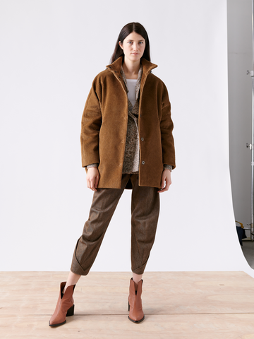 A woman stands on a wooden floor with a white backdrop, wearing the Long-Sleeved Bea Parka from Zero + Maria Cornejo, beautifully paired with gray pants and brown ankle boots.