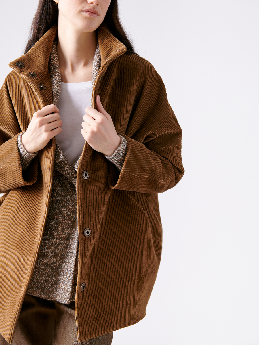 Wearing the Long-Sleeved Bea Parka by Zero + Maria Cornejo over a sweater and white shirt, a person faces forward adjusting the parka. The plain white background highlights the texture and style of their cotton fabric ensemble.