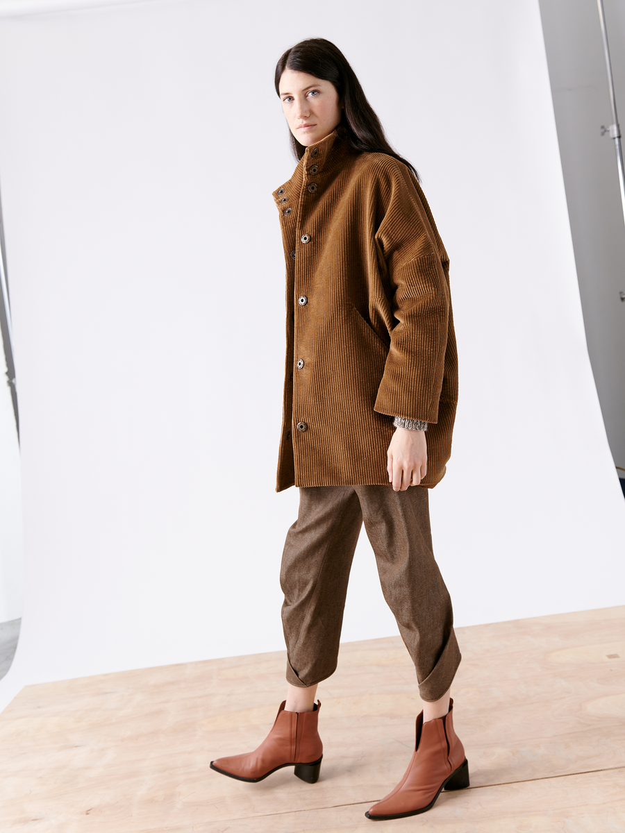 A person with long dark hair wears Zero + Maria Cornejo's Long-Sleeved Bea Parka paired with brown pants and ankle boots, posing against a plain studio backdrop. The draped parka enhances the casual, earthy vibe of the outfit.