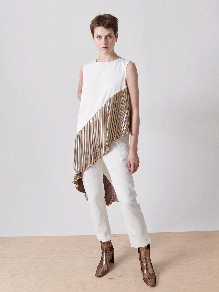 A person in the Zero + Maria Cornejo Plisse Heli Tunic, with one side metallic plissé, is paired with white pants and brown boots. They have short hair and stand on a light wooden floor against a plain backdrop.