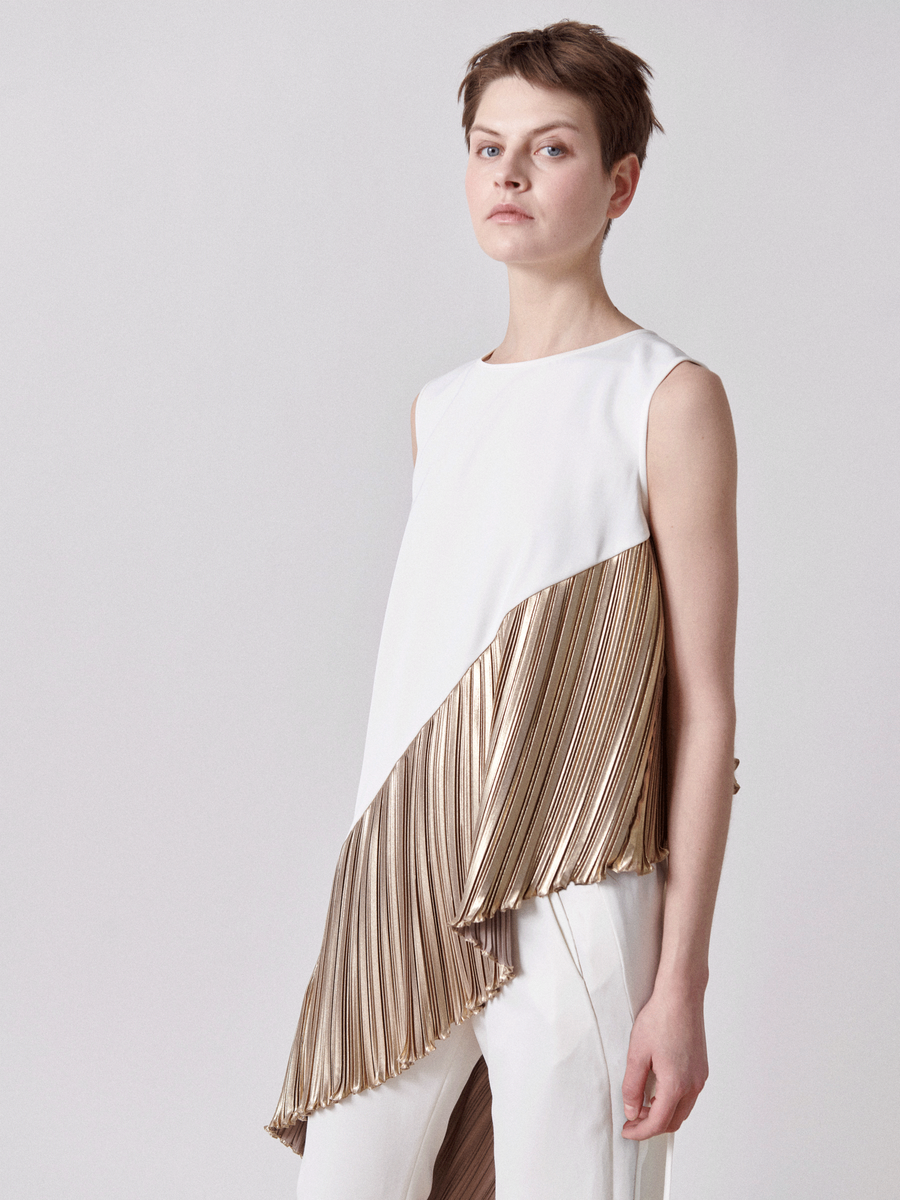 A short-haired person wears the Plisse Heli Tunic by Zero + Maria Cornejo, a white asymmetrical sleeveless tunic with diagonal metallic plissé, paired with white pants. They stand against a plain light gray background, looking slightly to the left.