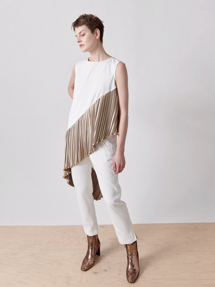 A person with short hair wears the Plisse Heli Tunic from Zero + Maria Cornejo, featuring an asymmetrical metallic plissé combo on one side. They pair it with white pants and brown snake-print boots on a wooden floor against a plain white background.