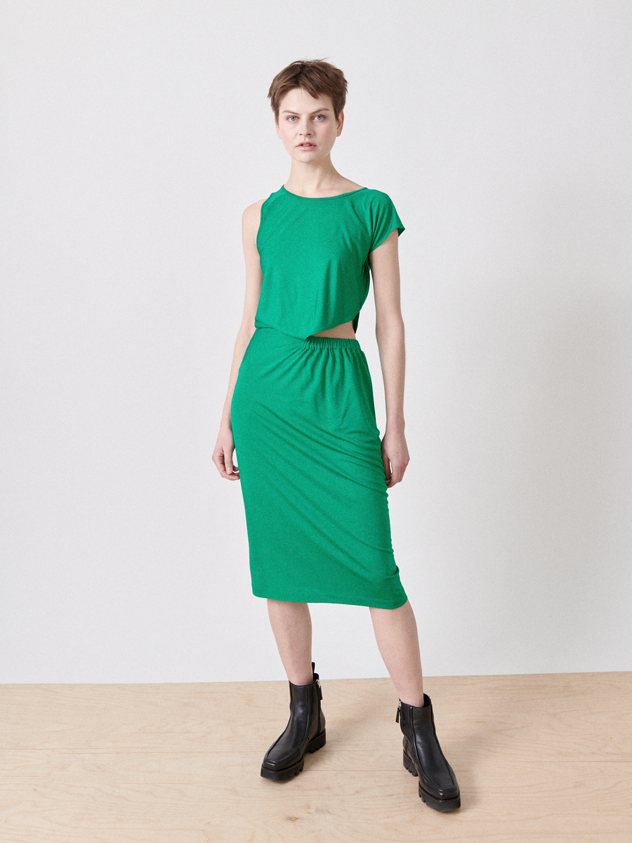 A person stands on a wooden floor against a plain background, wearing Zero + Maria Cornejo's Cut Out Triangle Dress, an asymmetrical sleeveless green dress with side cut-outs, paired with black ankle boots. They have short hair and gaze at the camera with a neutral expression.