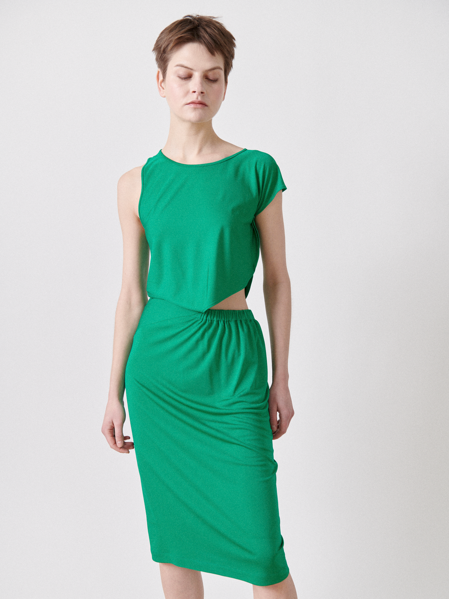 A person stands against a plain white background, wearing the Zero + Maria Cornejo Cut Out Triangle Dress - Final Sale, a lightweight sleeveless green dress with an asymmetrical waist cutout.