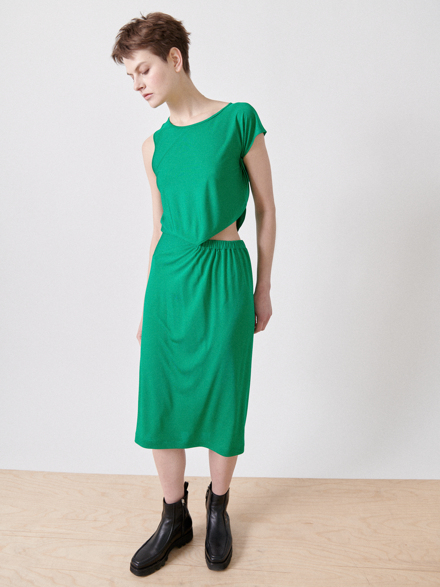 Someone with short hair wears the vibrant green Cut Out Triangle Dress by Zero + Maria Cornejo, featuring an asymmetrical triangle cut-out and an elastic banded waist. They pair it with black ankle boots, standing on a wooden floor against a plain white background, looking down to the side.