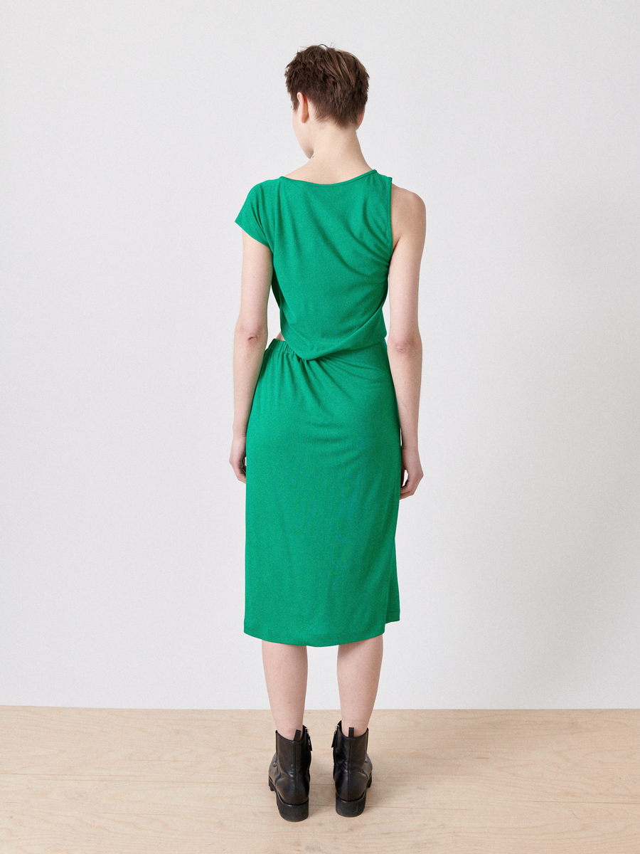 A person stands on a light wood floor against a white backdrop, wearing Zero + Maria Cornejo's Cut Out Triangle Dress in green. The knee-length dress features an asymmetrical back knot and cap sleeves, effortlessly paired with black ankle boots for modern elegance.