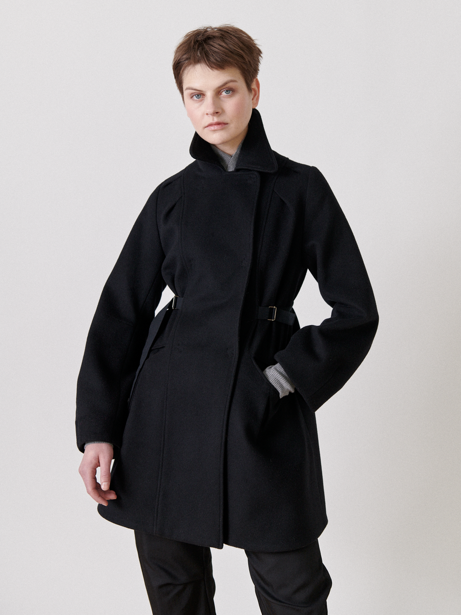 Against a plain white backdrop, a short-haired individual exudes confidence and style wearing the Zero + Maria Cornejo Leila Coat—a long black wool coat with an oversized collar—with dark pants, one hand casually in their pocket, looking at the camera.