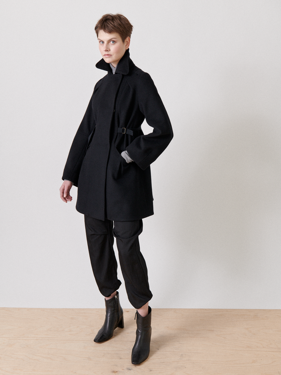 A person stands against a plain backdrop, wearing the Leila Coat by Zero + Maria Cornejo, featuring an oversized collar and grosgrain belt detail. They pair it with dark pants and black ankle boots, showcasing short hair and a neutral expression.