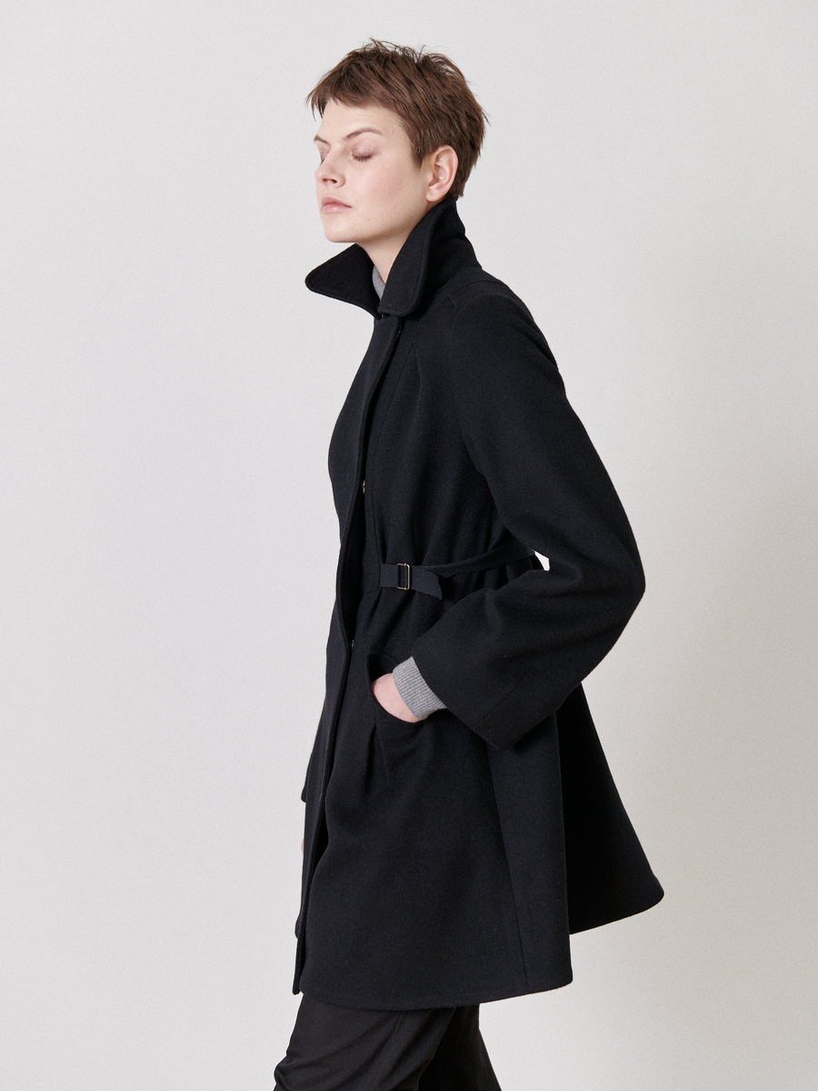 A person with short hair wears the Leila Coat from Zero + Maria Cornejo in profile, eyes closed. The black wool coat features an oversized collar and grosgrain belt detail, set against a plain, light-colored background.