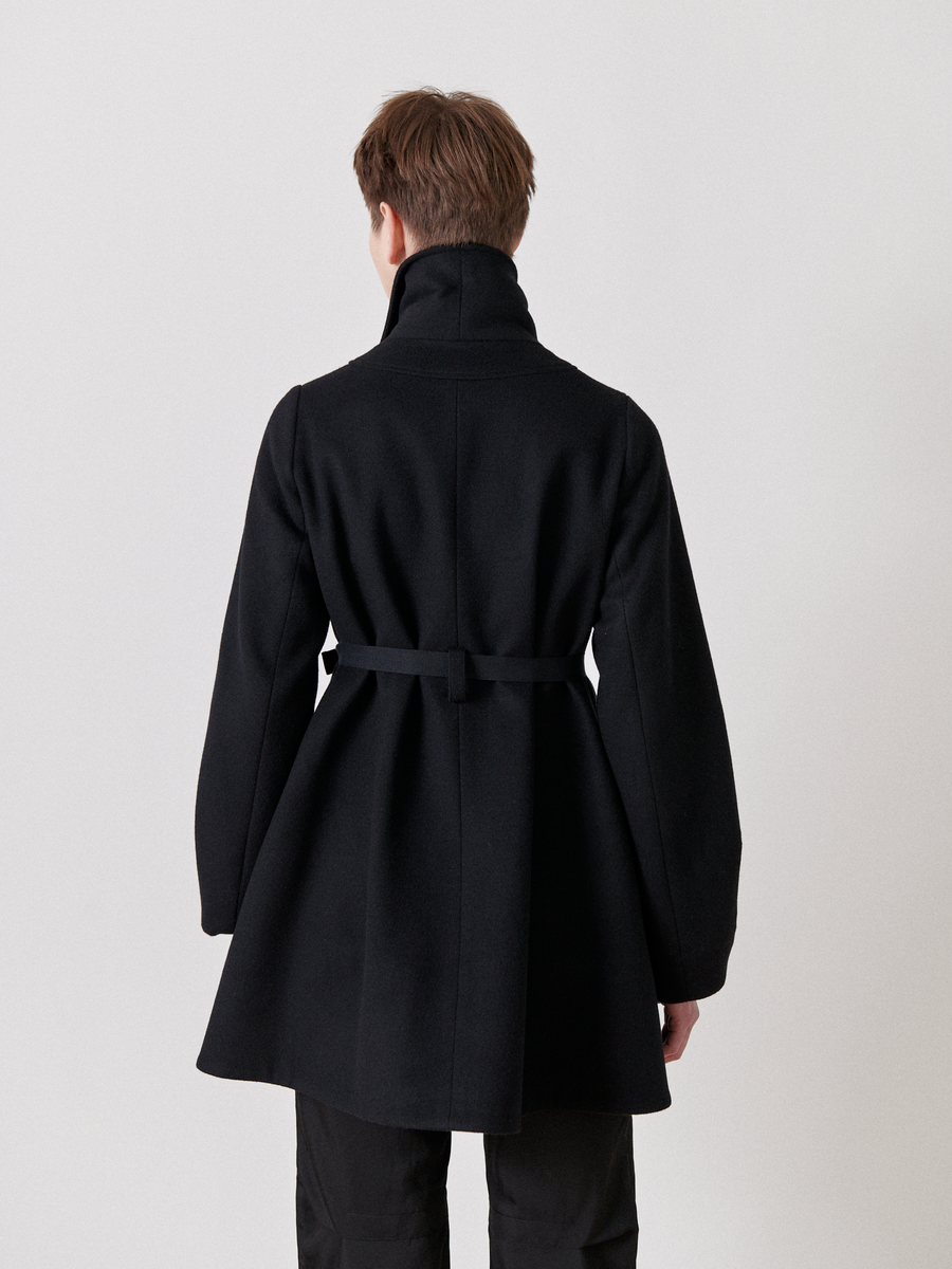 Seen from behind, a person with short brown hair wears the Leila Coat by Zero + Maria Cornejo. This black wool coat, featuring an oversized collar and a grosgrain belt at the waist, stands out against the plain white background.