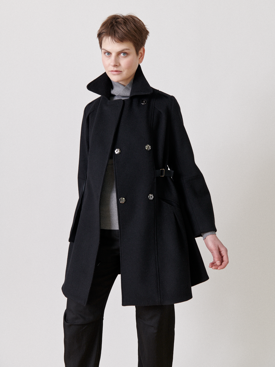 A person with short hair wears the Leila Coat, a black wool piece by Zero + Maria Cornejo, featuring an oversized collar and button details. It is styled with a grosgrain belt at the waist. They stand with a neutral expression against a plain light background.