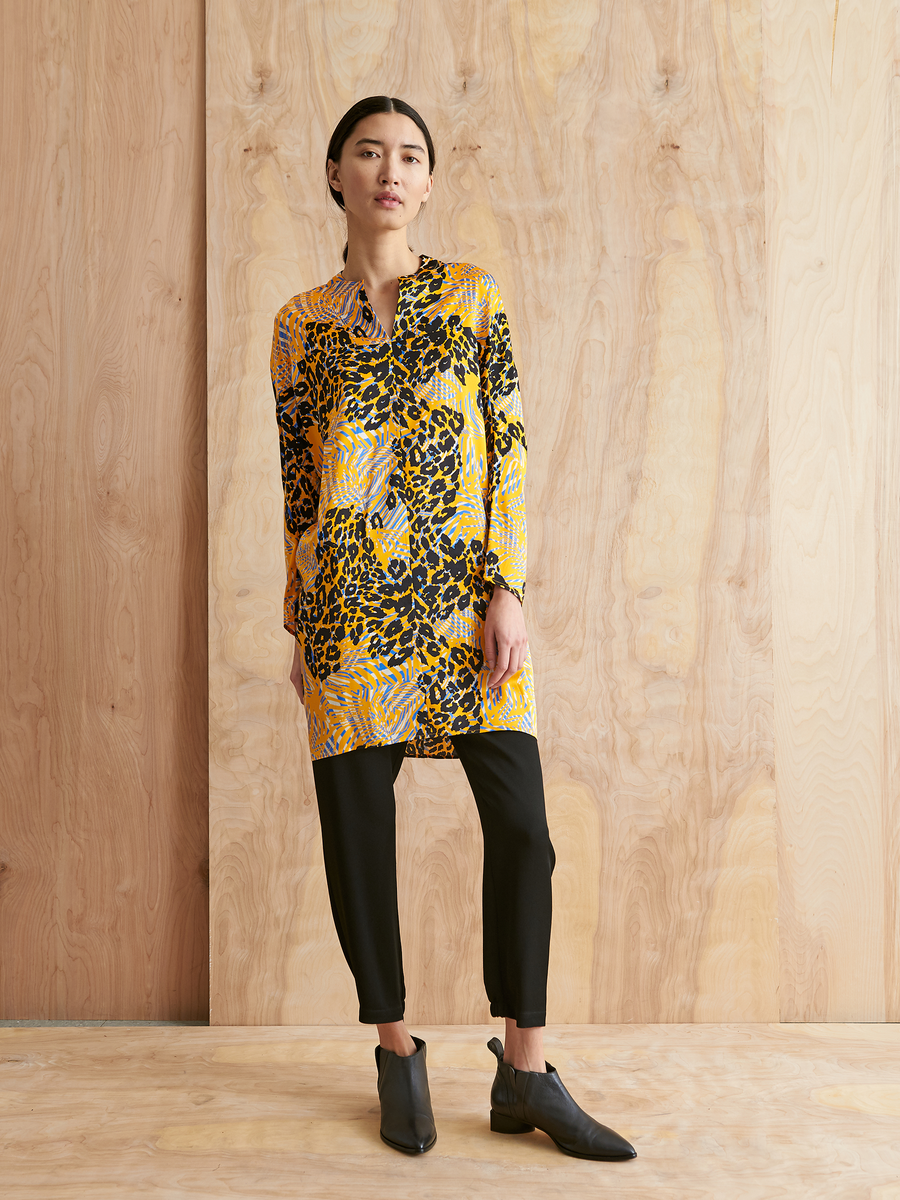 A woman stands against a wooden background, confidently wearing the Zero + Maria Cornejo Long-Sleeved Hex Tunic Dress with black slim pants and ankle boots. Her straight dark hair is pulled back. Made in New York.