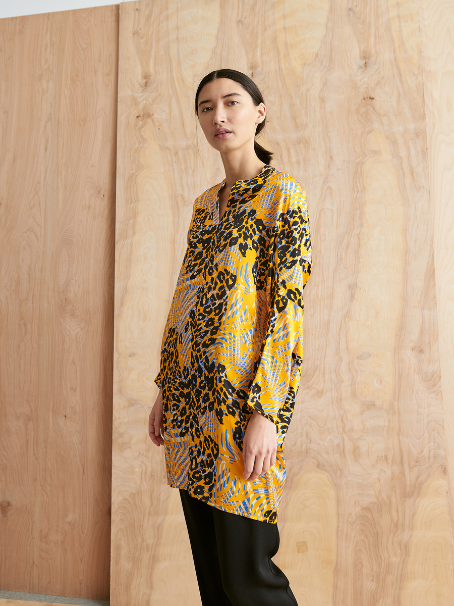 A woman with long dark hair wears the Zero + Maria Cornejo Long-Sleeved Hex Tunic Dress with black pants, standing in front of a light wooden wall.