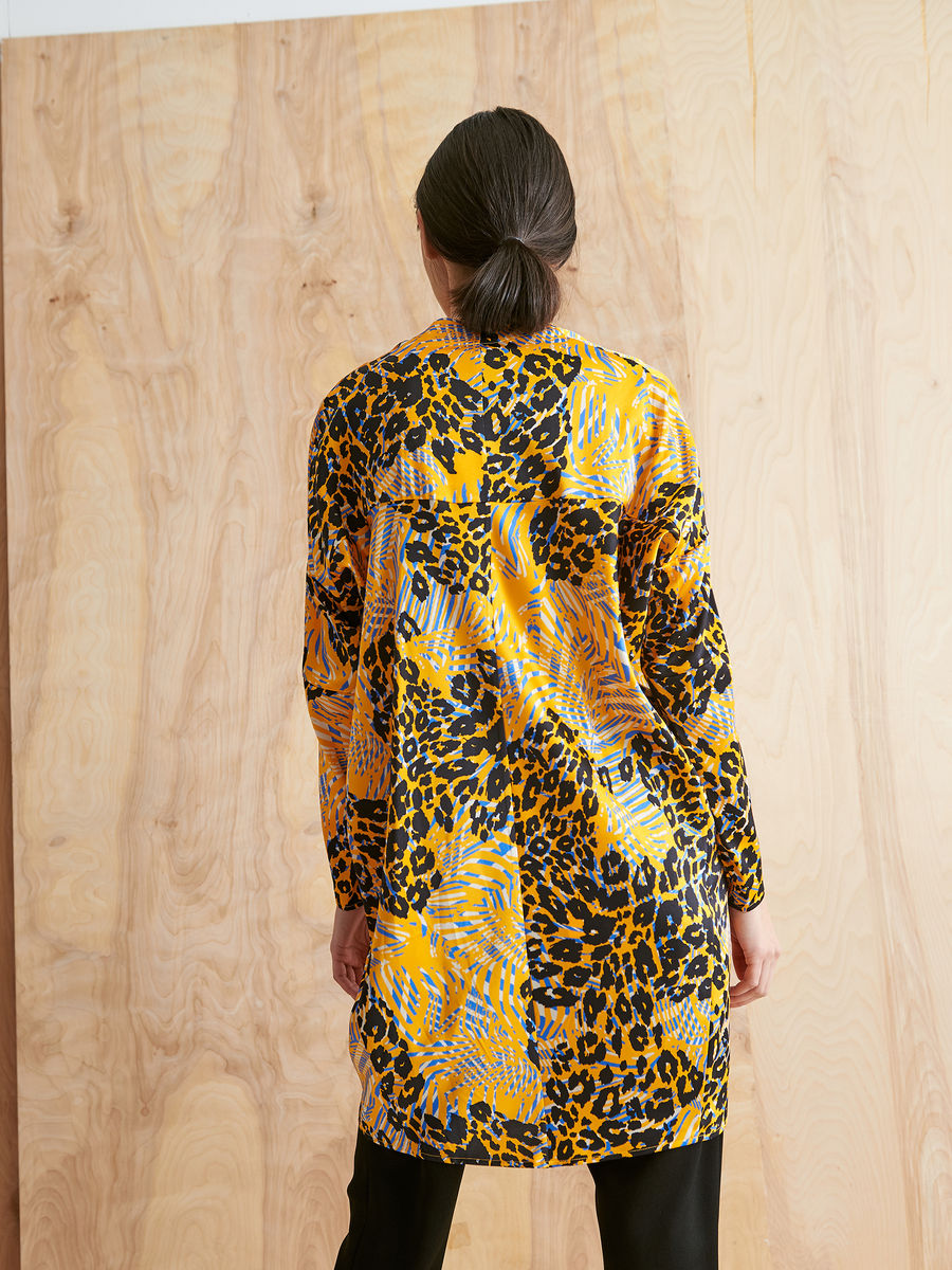 A person with dark hair in a low bun faces away, wearing the Zero + Maria Cornejo Long-Sleeved Hex Tunic Dress in cheetah print with black pants, set against a light wooden background.