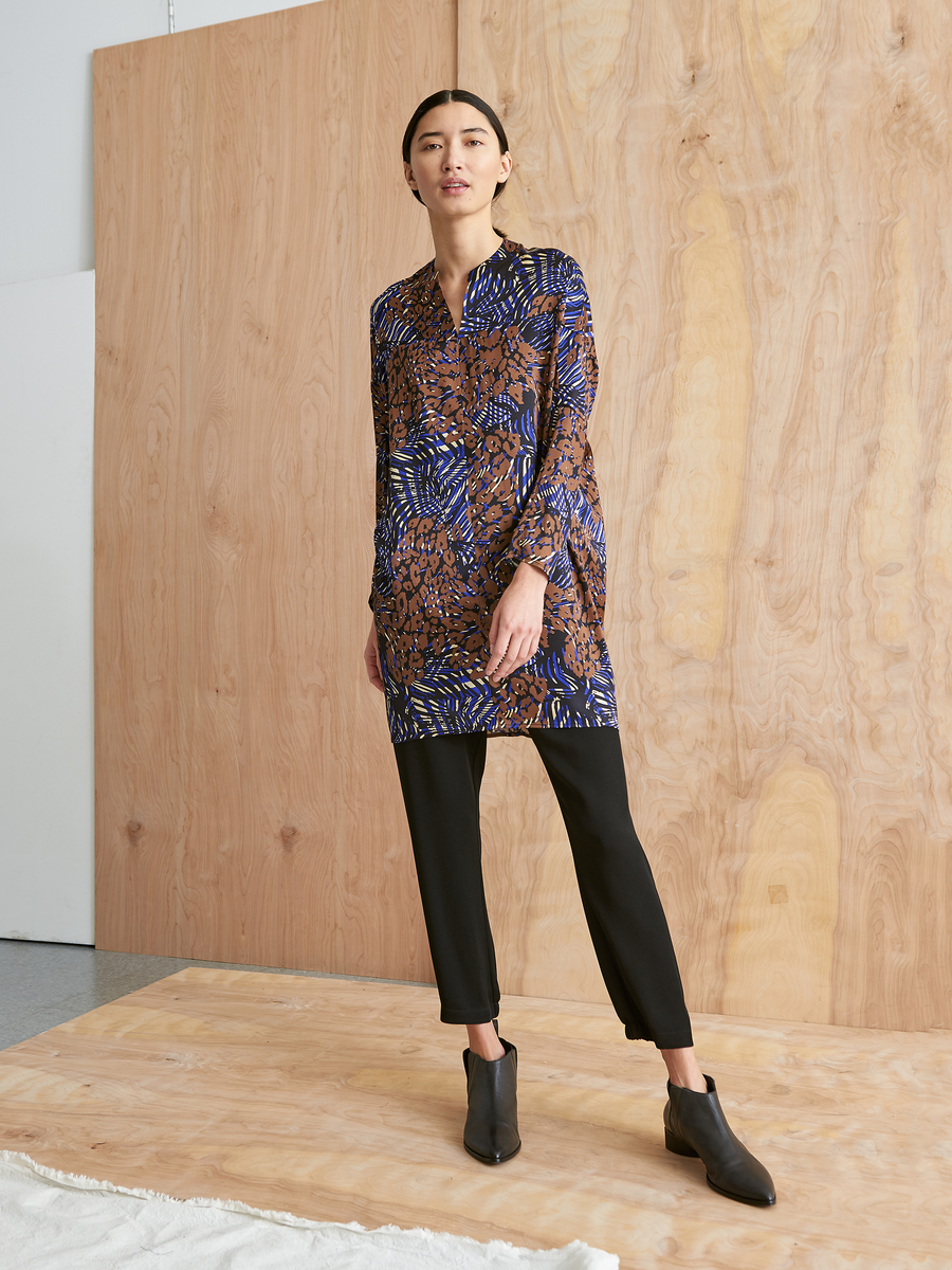 A woman stands indoors on a wooden floor against a wood-paneled wall, wearing the Zero + Maria Cornejo Long-Sleeved Hex Tunic Dress with brown and blue cheetah print, paired with black pants and black ankle boots.